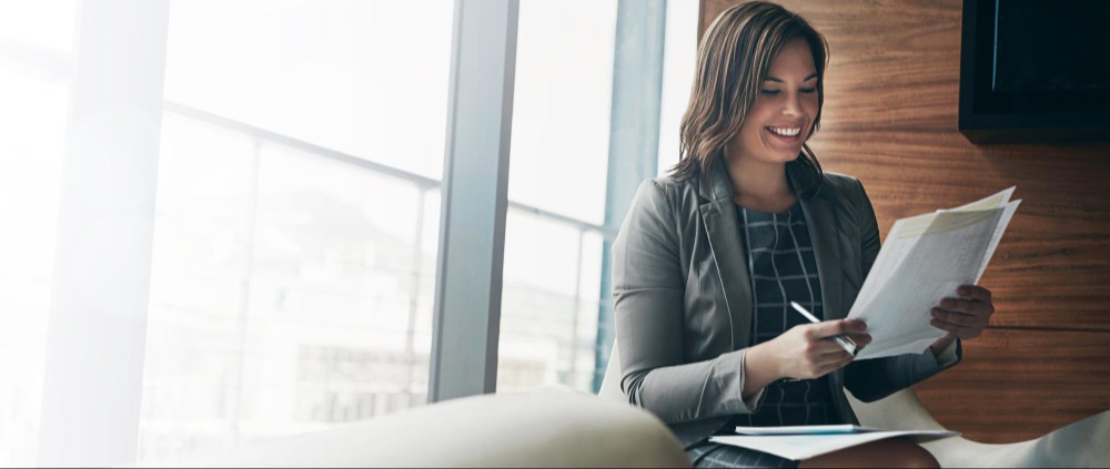 Confident female entrepreneur smiling while reading paperwork in a bright, modern corporate setting.