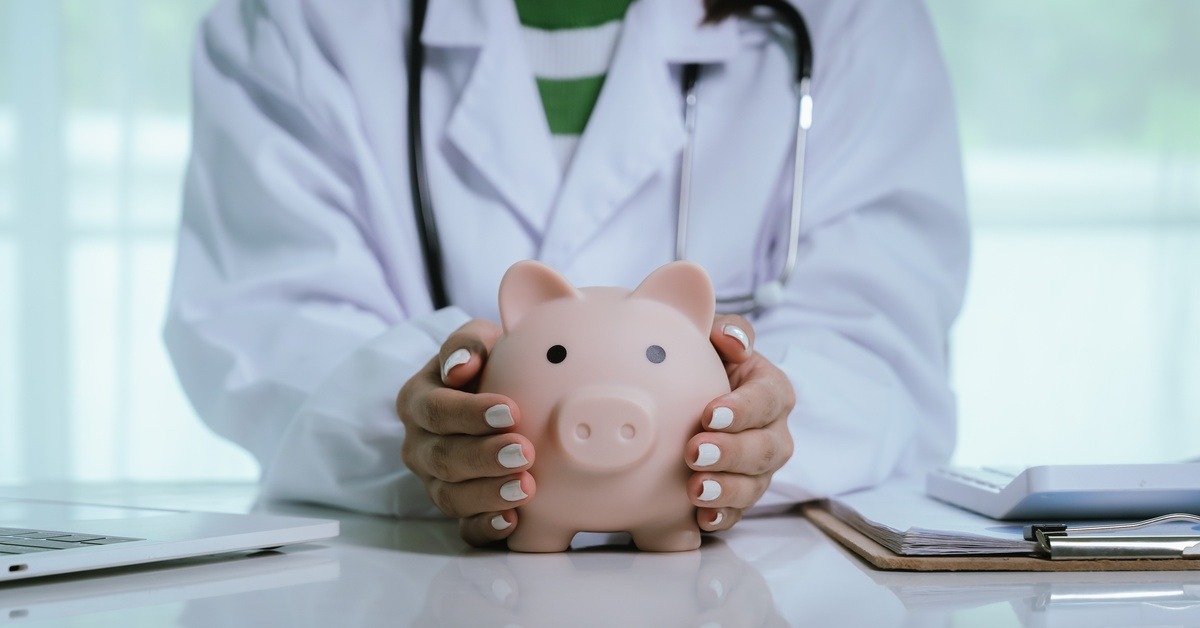 A physician wearing a white lab coat stands behind a desk and cups the sides of a large piggy bank with her manicured hands.