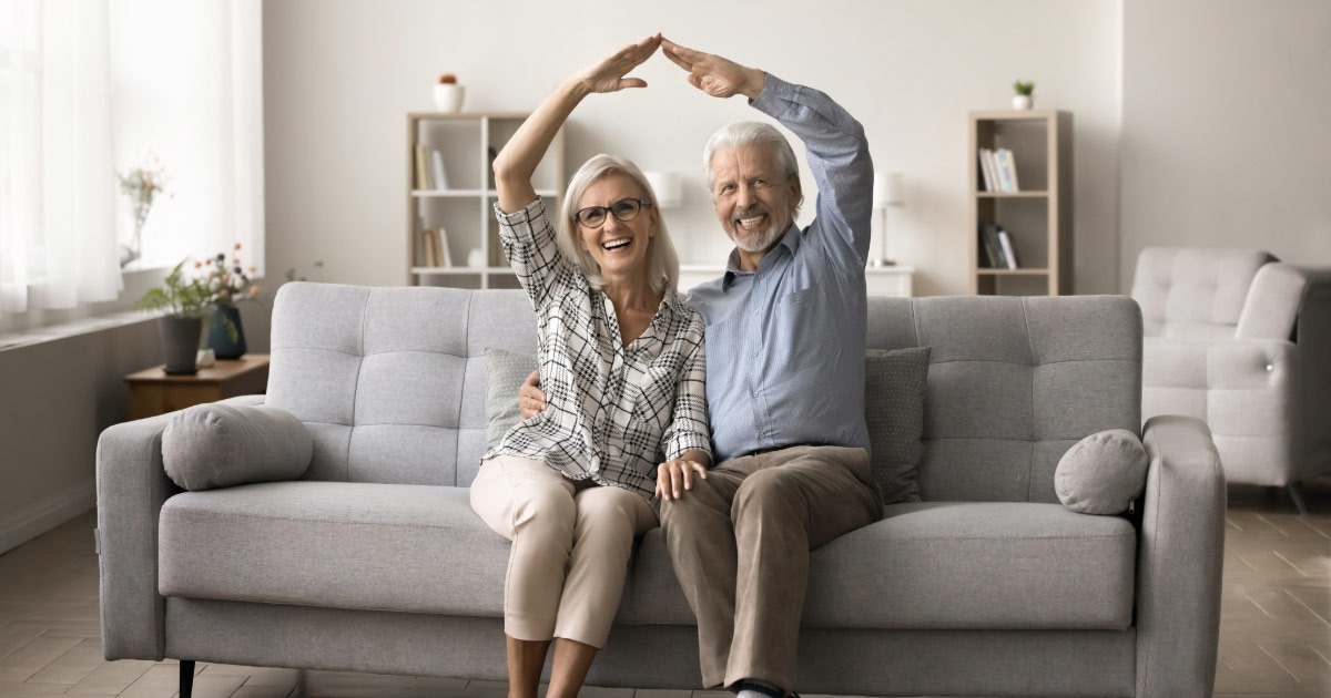 Older couple making a house shape with their arms while sitting on couch