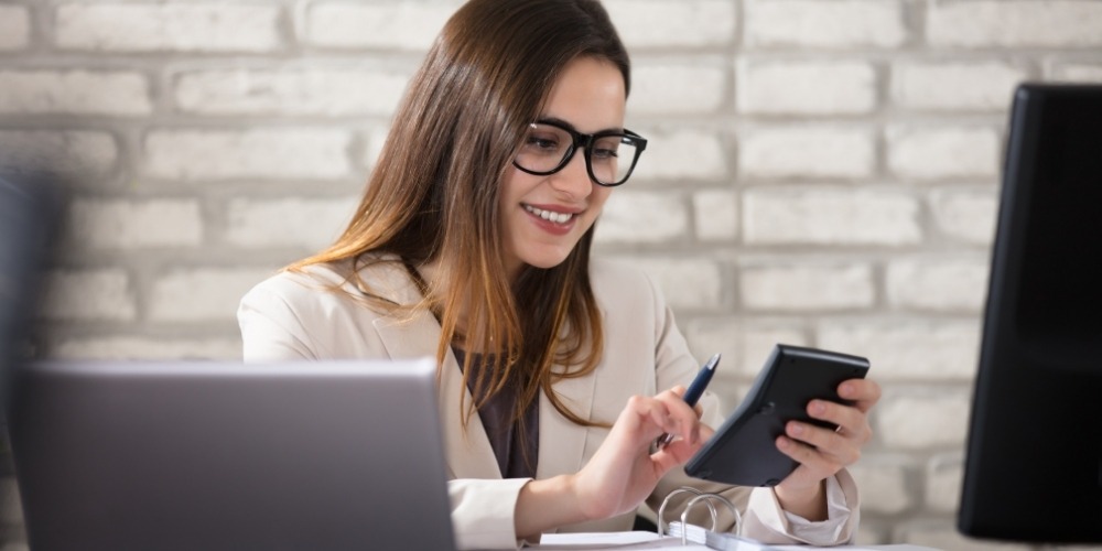woman with glasses sitting at an office desk uses a handheld calculator