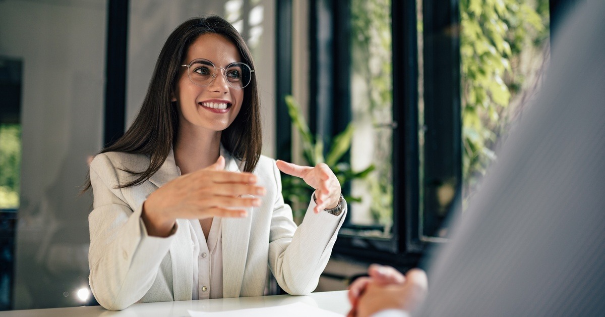 A professionally dressed woman smiles and gestures with her hands as she talks to a client sitting across from her.