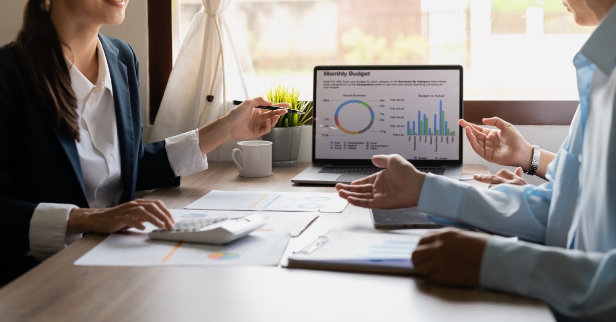 A professionally dressed woman sitting across from two clients; she points at a tablet screen with graphs on it.