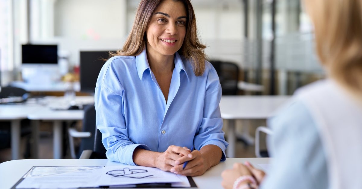 A woman in a blue blouse with her hands clasped on the desk, smiling at the person across from her.