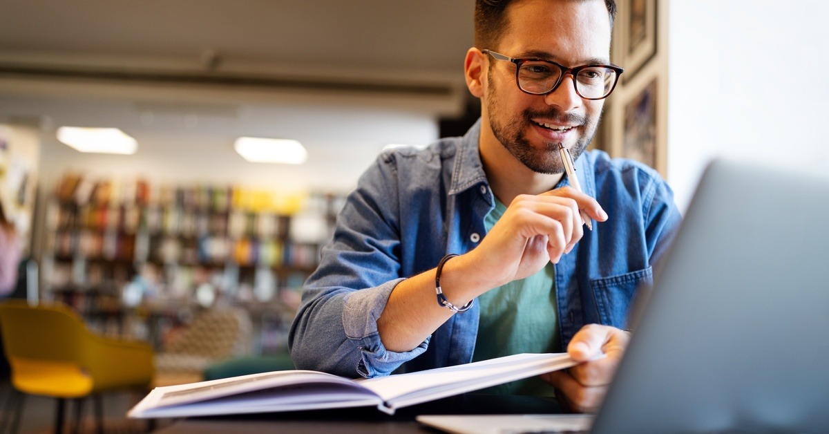 A bearded student taps a pen to his chin as he studies in a library from a laptop and an open book.
