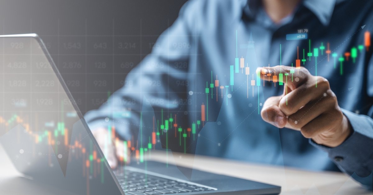 A man in a blue button-up shirt, sitting at a desk, pointing his finger at a trade graph in the foreground.