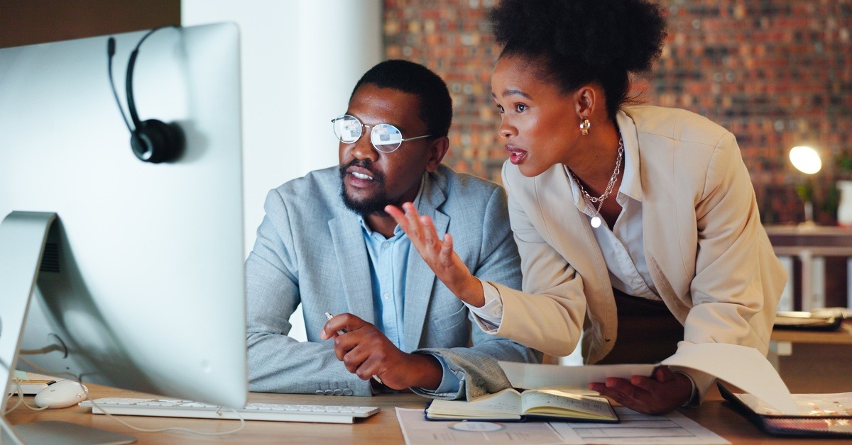 Two professionally dressed people; the man in glasses sits at his laptop while the woman at his shoulder gestures in confusion.