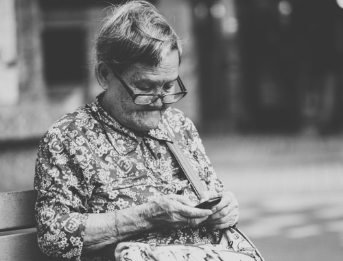 An older woman sitting on a bench looking at her phone, in black and white, illustrating financial planning for widows.