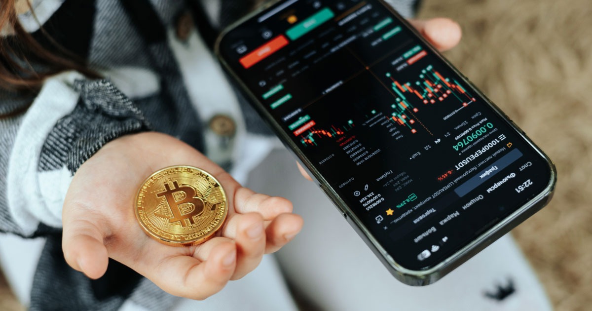 Man holding gold Bitcoin in his hand while checking stocks on his phone