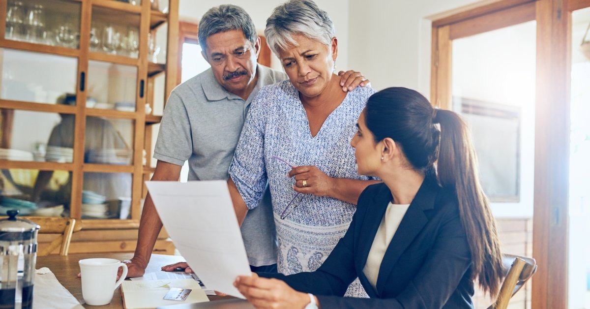 An older couple is standing and looking down at a professionally dressed woman holding up a piece of paper.