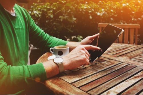 man using a tablet and drinking coffee at an outdoor picnic table