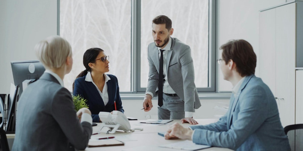 a corporate meeting in a bright office, where a man in a grey suit stands at the head of a conference table