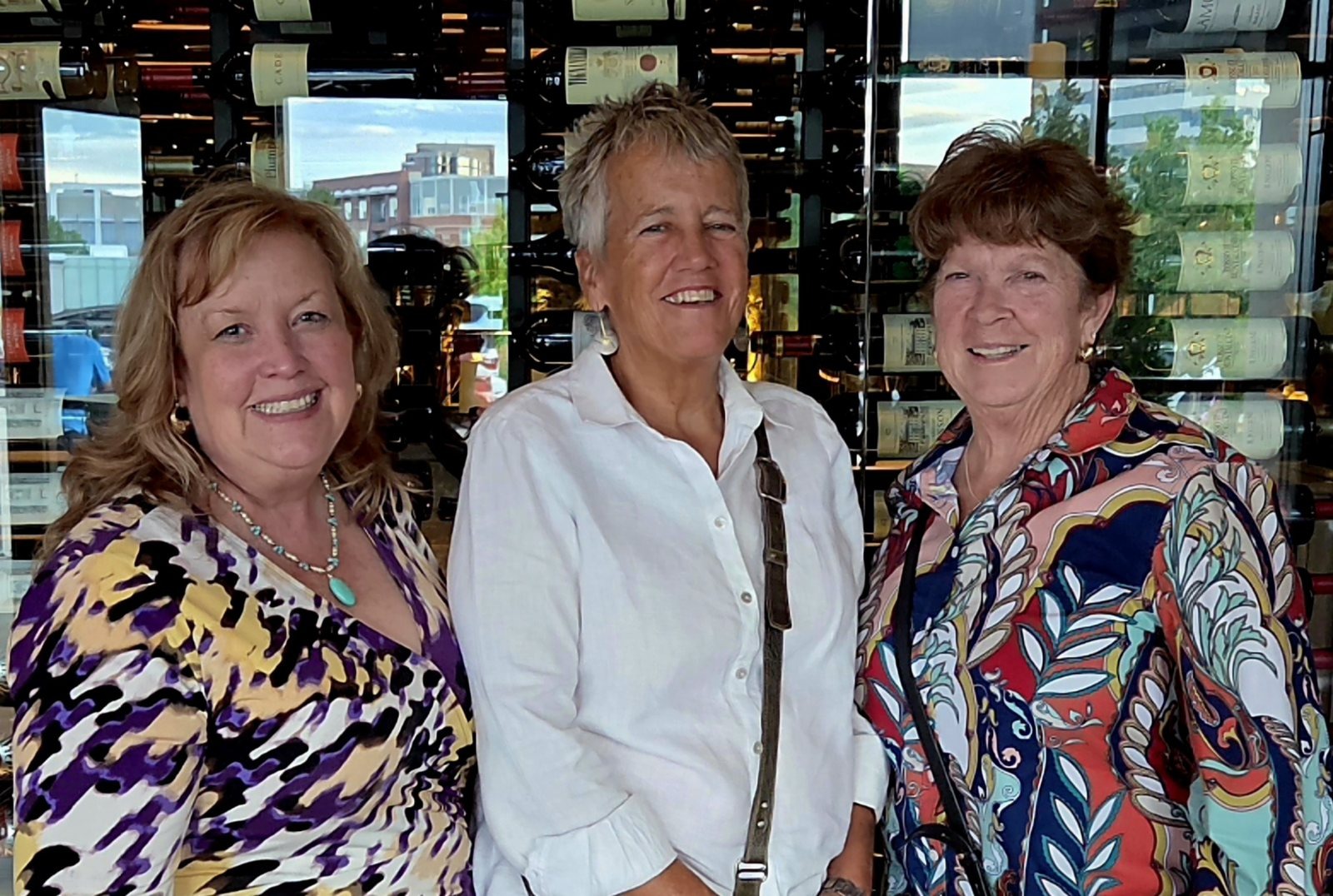 Three women standing together in front of a floor-to-ceiling wine rack