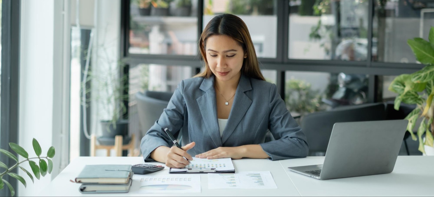 Businesswoman recording financial data and reviewing bookkeeping documents with a calculator and laptop.