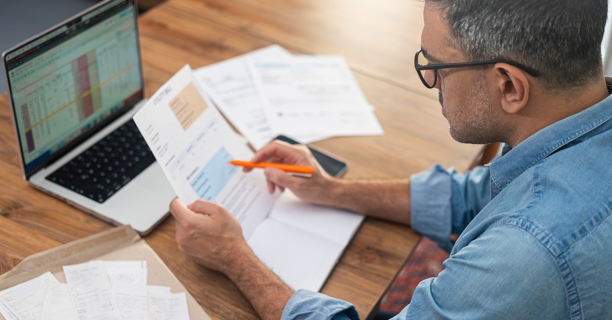 An over-the-shoulder shot of a man sitting at his laptop reviewing paperwork with an orange writing implement.