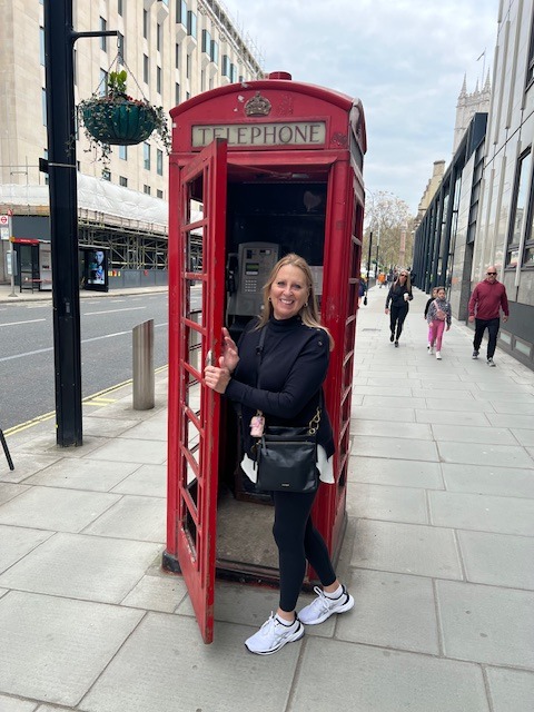Deborah stands before a bright red telephone booth, embodying a moment of nostalgia in a bustling urban environment.