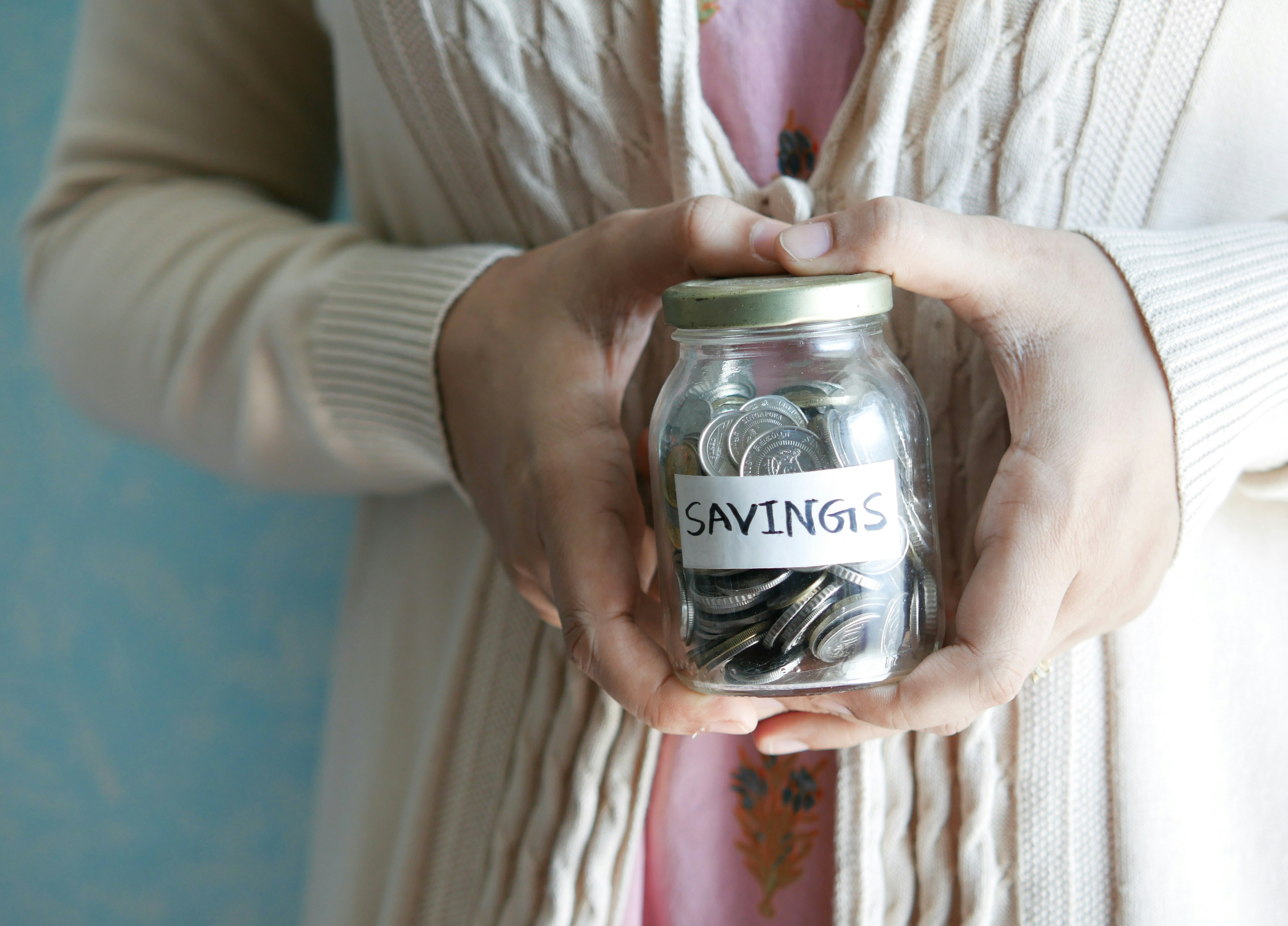 Hands holding a glass jar labeled “Savings” filled with coins ``