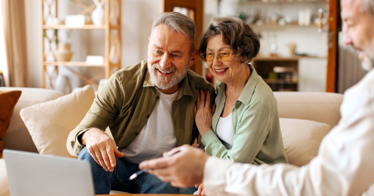 A happy older couple on a living room couch, looking at a laptop as their advisor gestures to the screen.