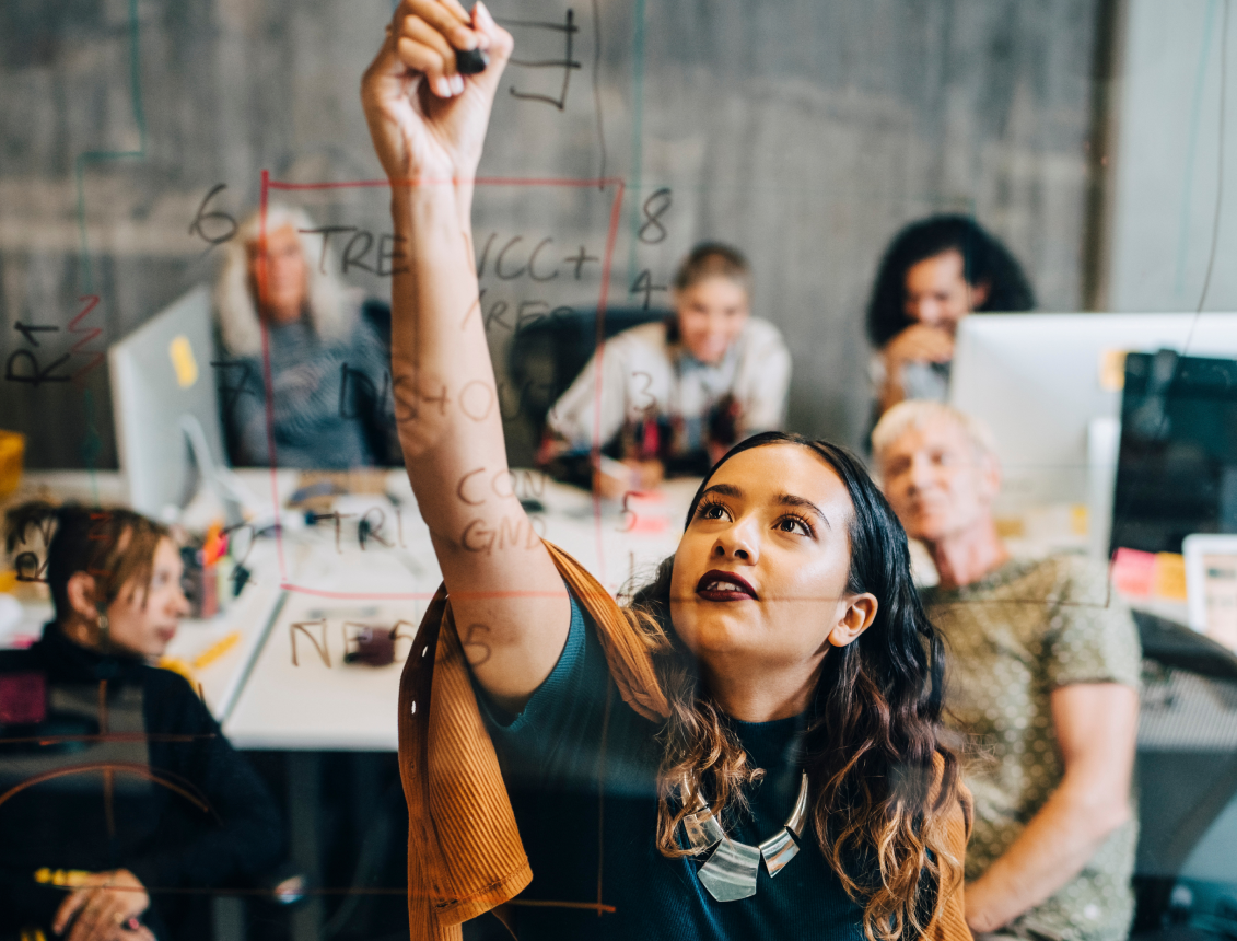 person doing a calculation on a transparent white board while others at a meeting table observe