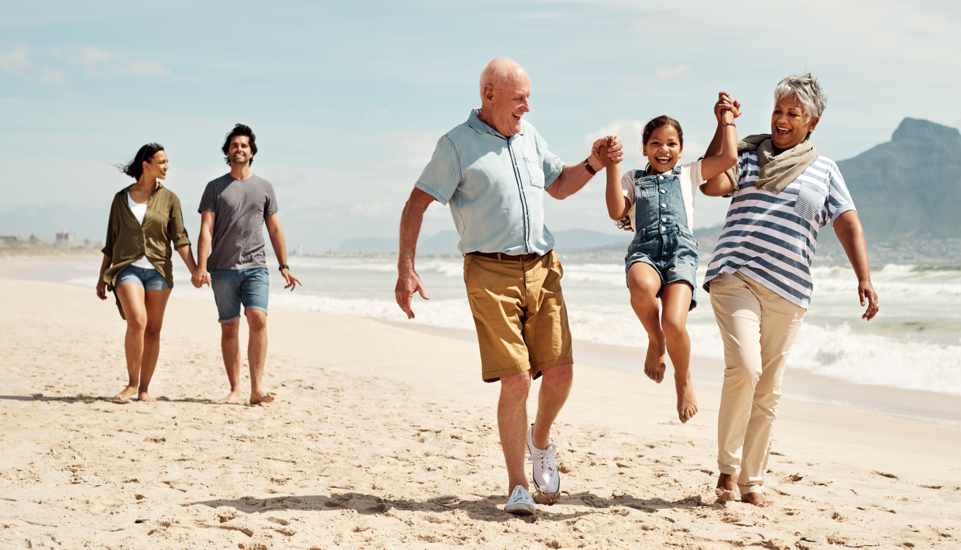 grandparents playing with their daughter on the beach and behind them is a couple holding hands