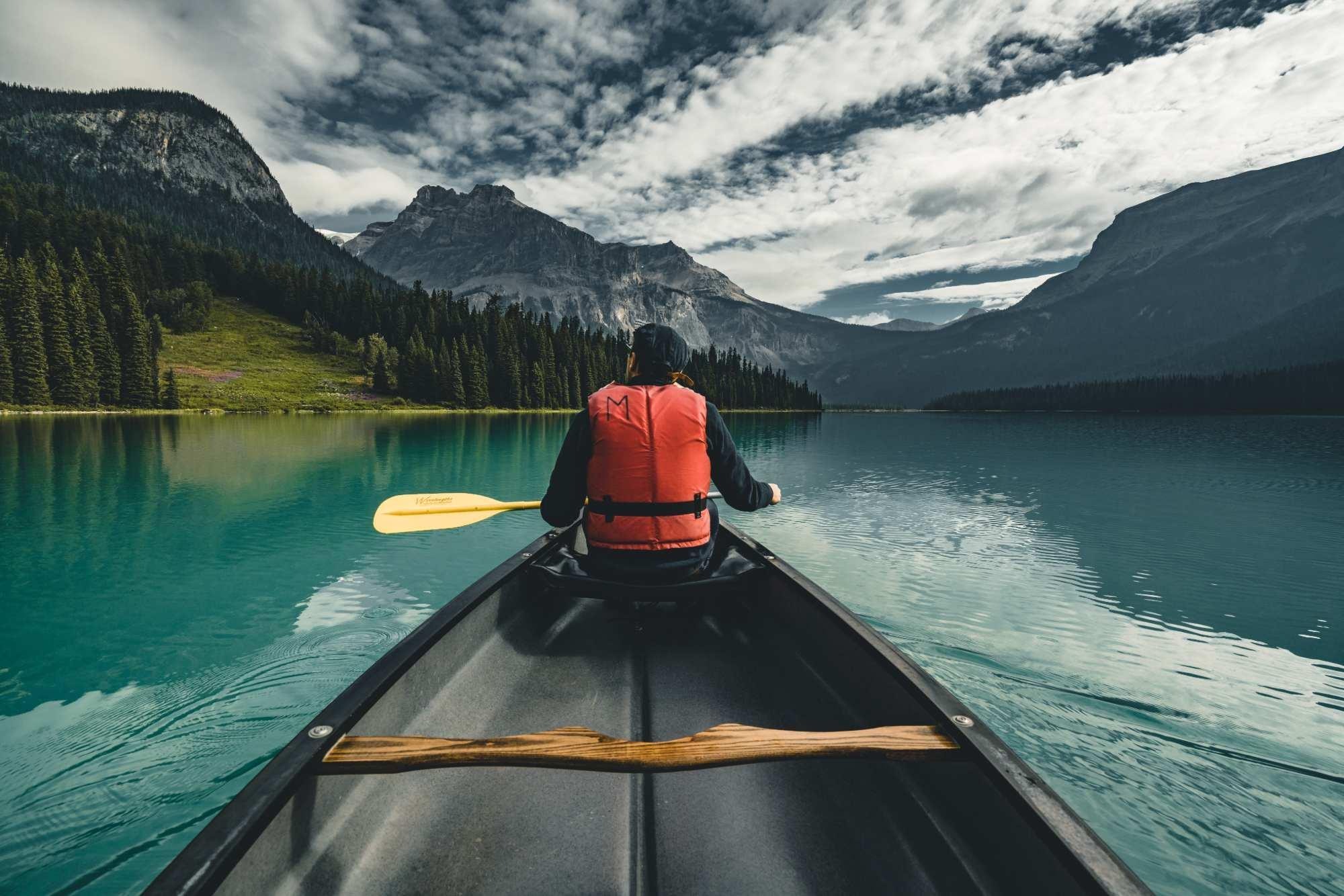 person sitting at the front of a canoe in water, looking at high mountains in the distance