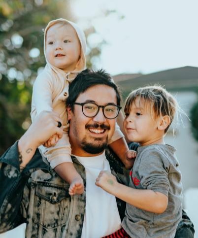 Man with glasses holding two small children outside