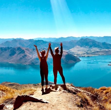 Two women at the top of a hike with there arms up overlooking a large lake with mountains