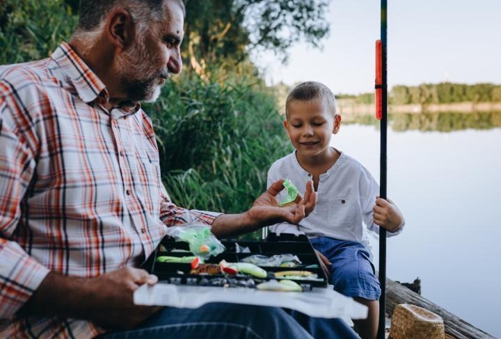 A man hands a fishing lure to a young boy next to a pond.