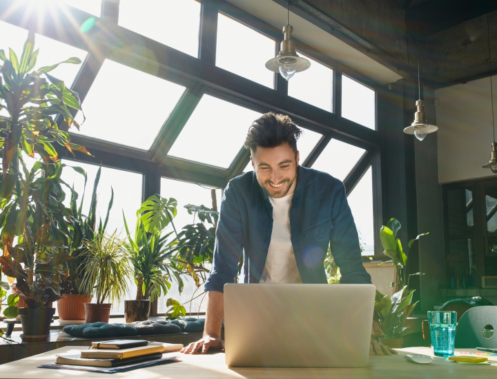 man in a sunny office with plants standing in front of a laptop and smiling