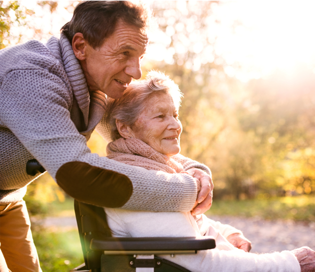 Photo of elderly man hugging an elderly woman sitting in a chair outside