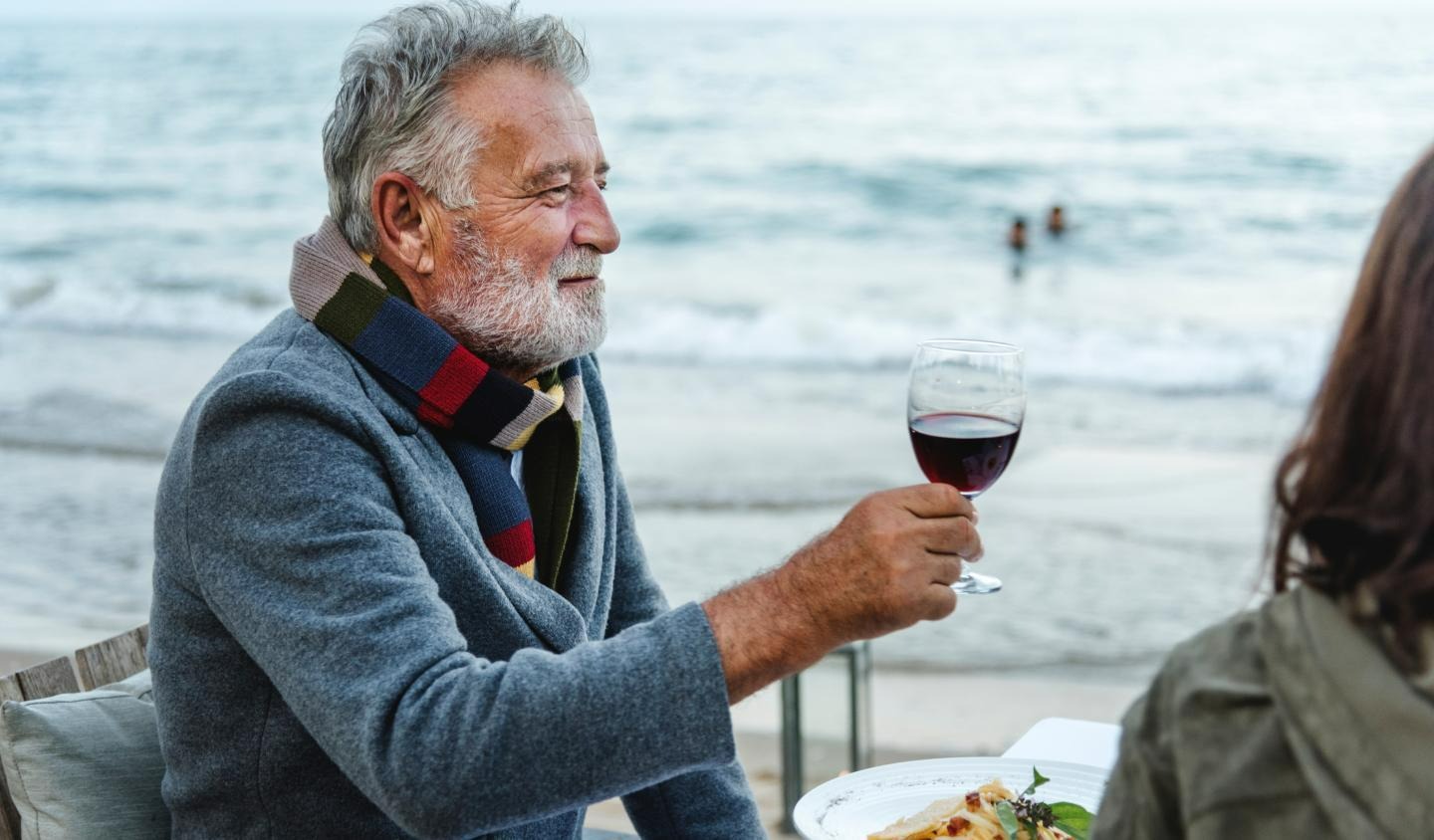 man eating a meal and drinking a glass of wine next to the ocean with a companion