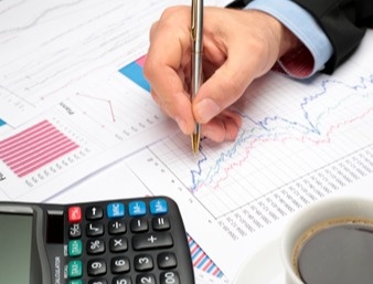 person writing on a document with graphs, using a calculator and drinking coffee