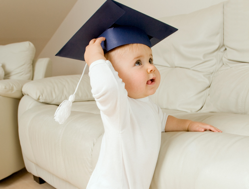 toddler holding on to a couch and wearing a graduation cap