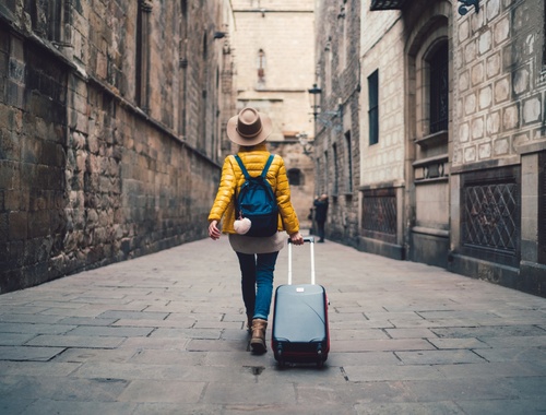 a woman dressed for travel and pulling a suitcase walking away from the camera down a narrow alleyway