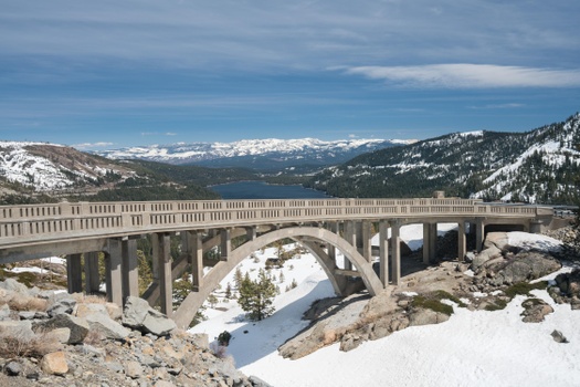 Bridge with snow underneath it, mountains and a lake in the distance
